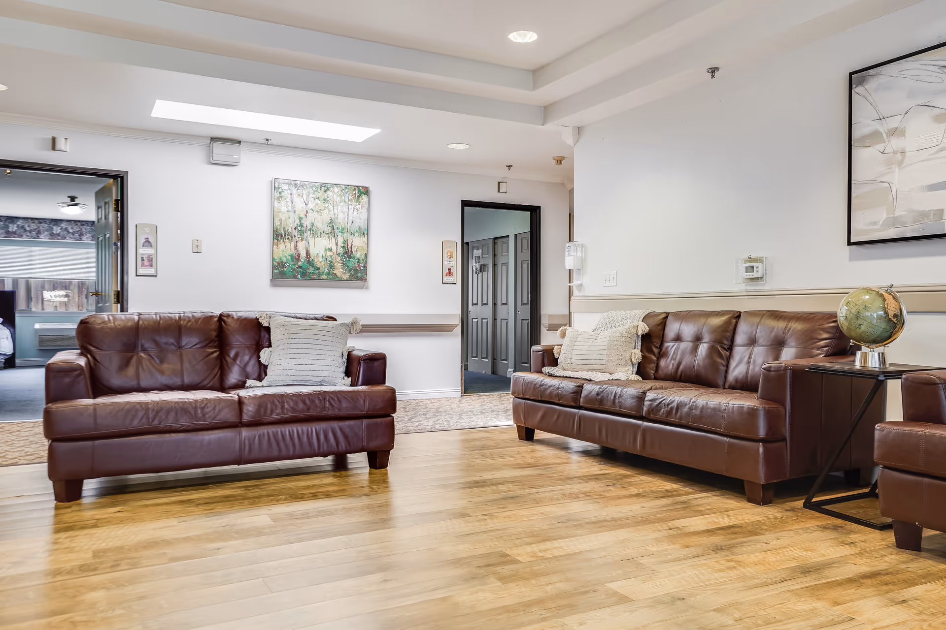 A senior living facility common area with two brown leather sofas facing each other, each with a decorative pillow. The room has light wooden flooring, white walls with artwork, and a skylight. There is a small side table with a globe on it next to one sofa. Two doorways lead to other rooms, one showing a bedroom with a bed and window.