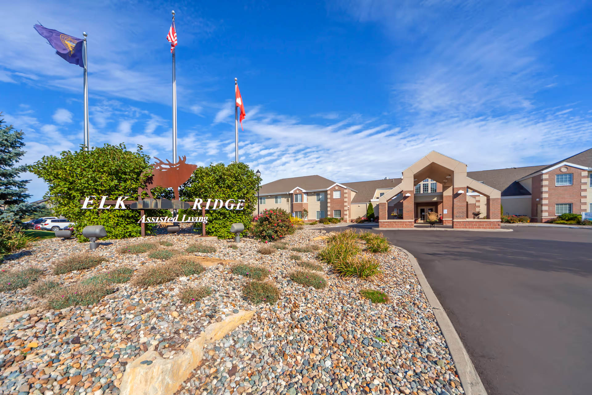 Exterior view of Elk Ridge Village Assisted Living facility showing the entrance with a covered drop-off area, a landscaped rock garden with shrubs, and a metal sign featuring an elk silhouette and the text 'Elk Ridge Assisted Living'. Three flagpoles with flags are visible behind the sign under a blue sky with scattered clouds.