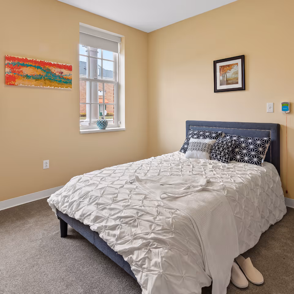 A neatly made bed with a white textured comforter and several decorative pillows in a bedroom with beige walls. There is a window with a white blind, a colorful abstract painting on one wall, and a framed picture of trees on another wall. A pair of slippers is placed on the carpeted floor next to the bed.