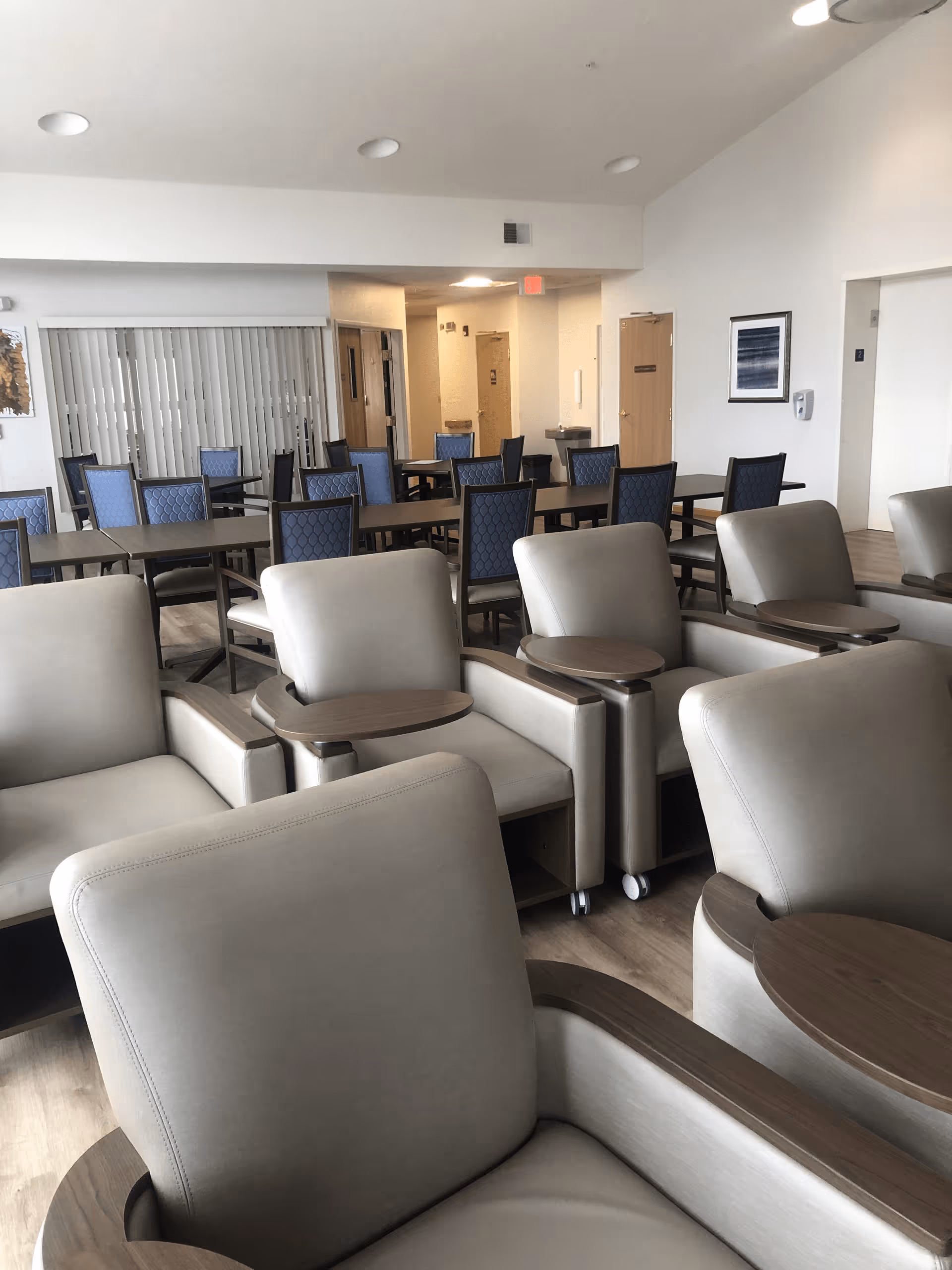 Bright communal lounge and dining area with rows of beige armchairs with attached wooden trays and tables with blue-upholstered dining chairs.