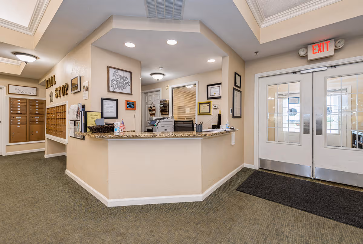 Reception area inside Christina House facility with a front desk featuring a granite countertop, office equipment, and framed certificates on the walls. To the left, there are mailboxes labeled 'Mail Stop' and a hallway. Double glass doors with an exit sign are visible on the right side.