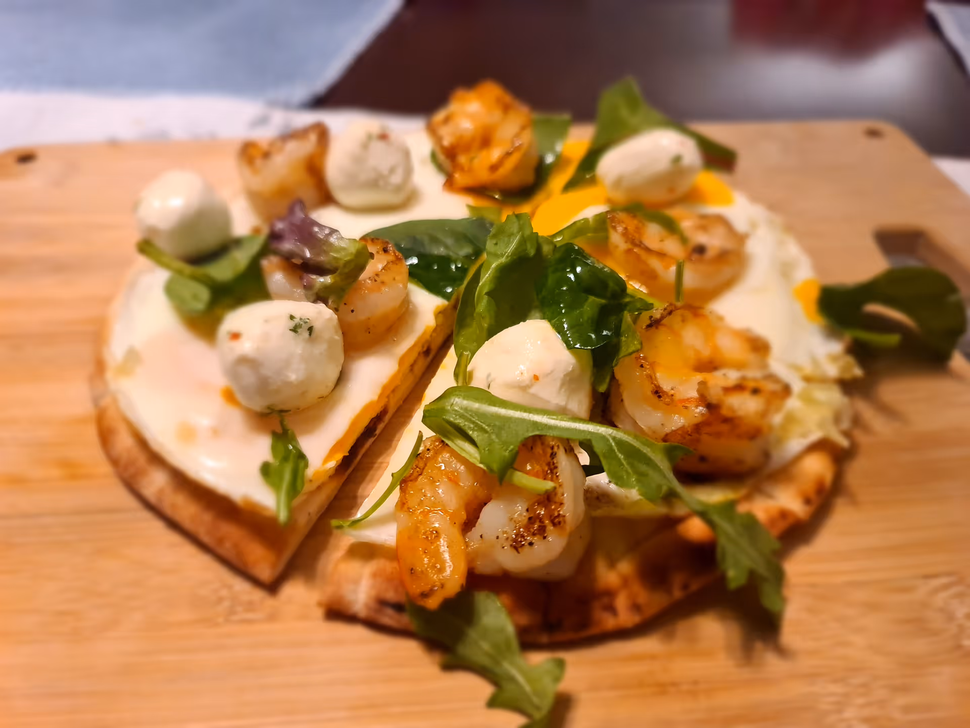 Grilled shrimp and small mozzarella balls with greens on a flatbread served on a wooden cutting board.