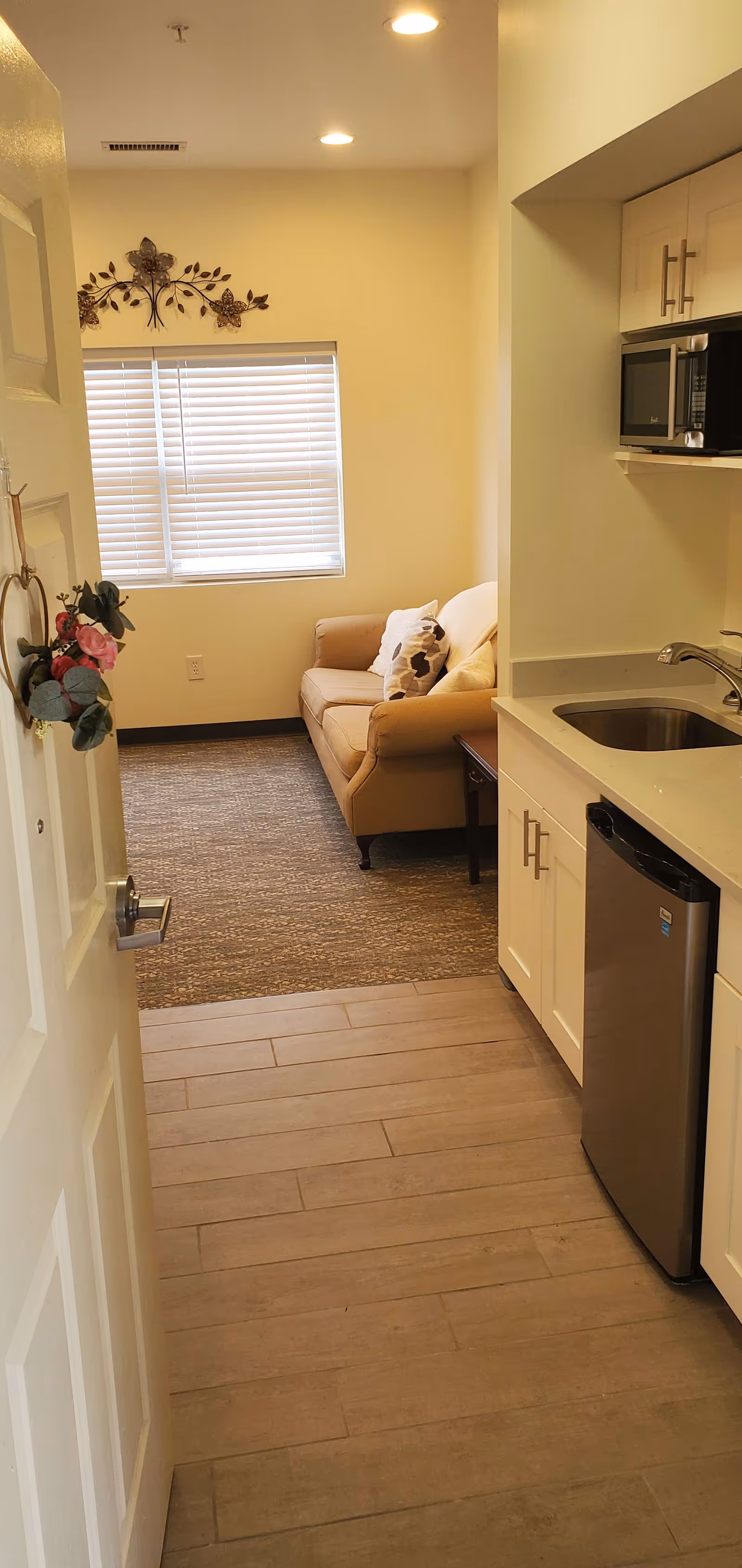 View down a small apartment entry showing a kitchenette with a sink and mini fridge on the right and a couch by a window at the end.