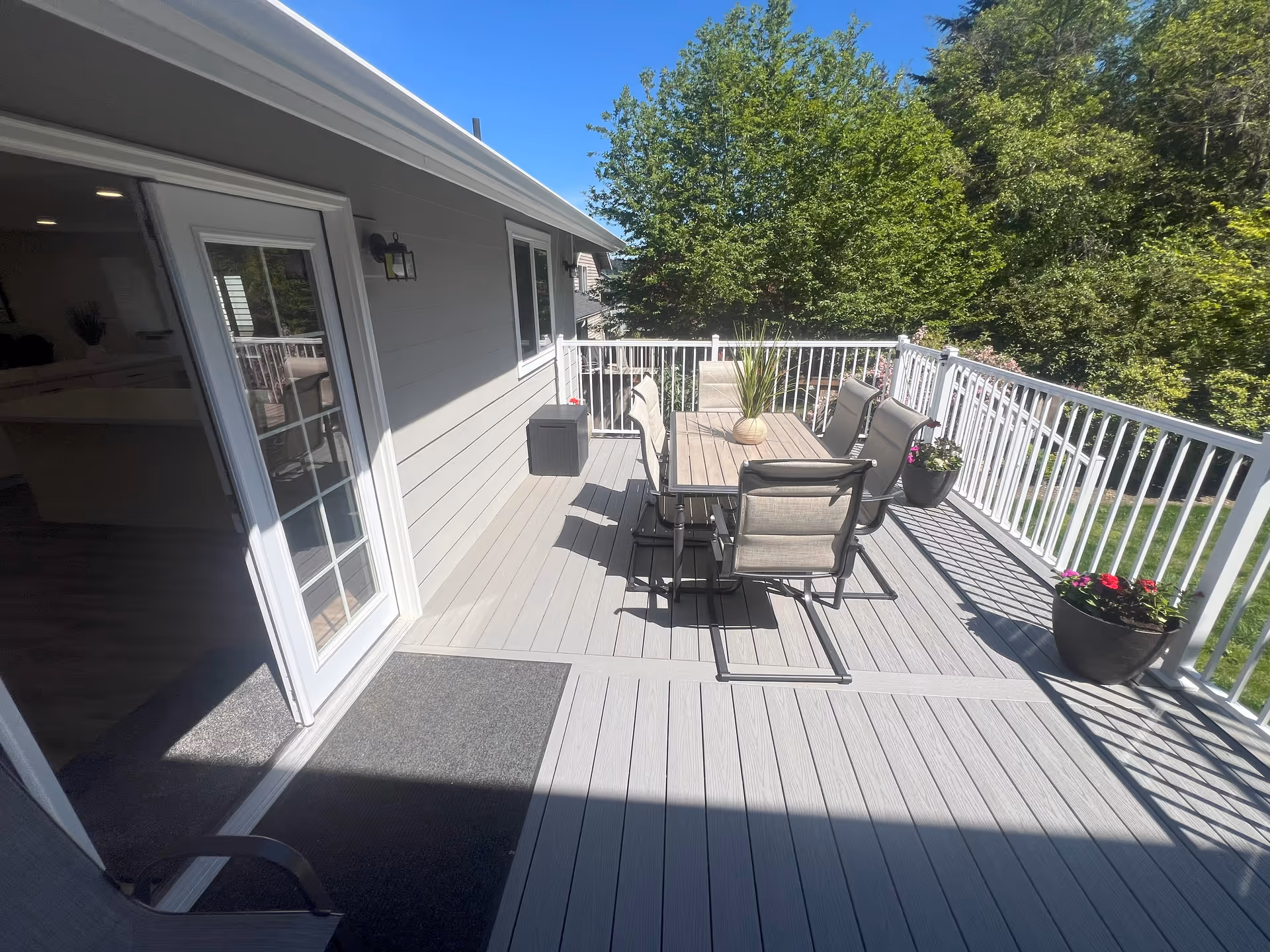Sunlit outdoor deck with a dining table and chairs, potted plants, white railing, and a sliding glass door leading inside.