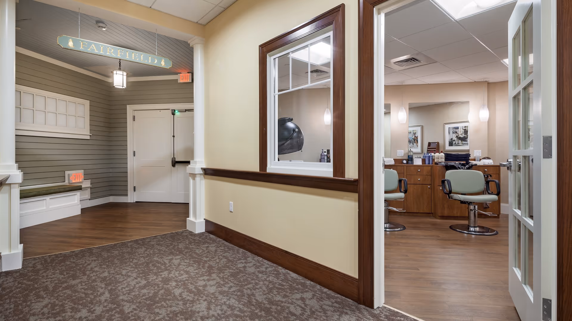 Interior hallway of a senior living facility with a sign reading 'Fairfield' hanging from the ceiling. The hallway has beige walls, wood trim, and a carpeted floor leading to a room with two green salon chairs, a large mirror, and wooden cabinets, suggesting a hair salon or grooming area.