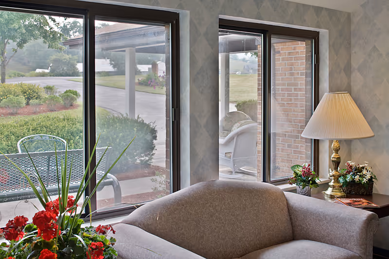 A cozy seating area with a sofa, side table with a lamp and flowers, and large windows looking out to a patio and landscaped grounds.