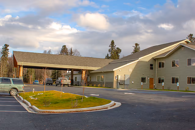 Front exterior of a senior living facility with a covered entryway, parking lot, and lawn under a partly cloudy sky.