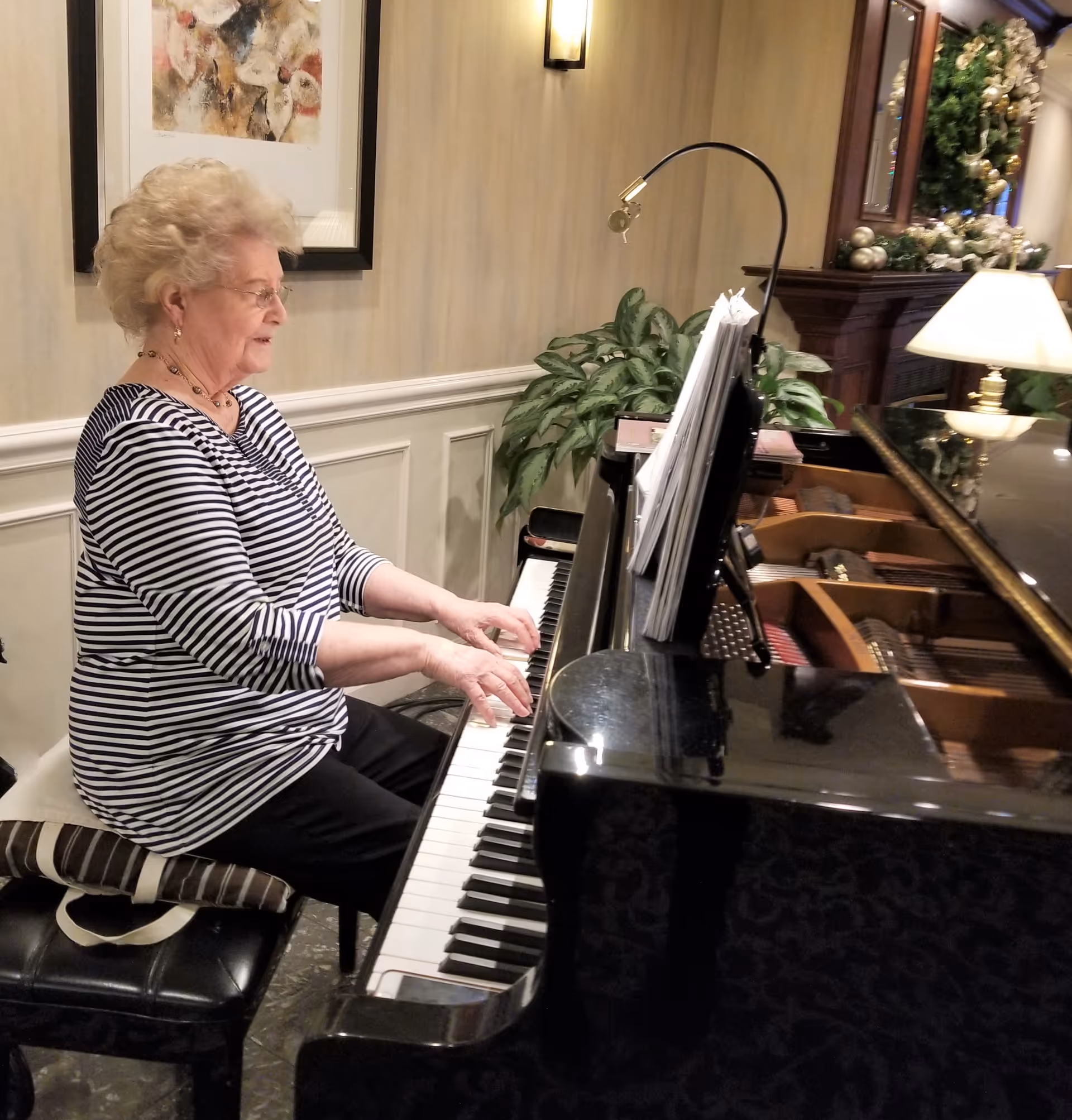 An elderly woman with curly white hair and glasses is playing a grand piano in a warmly decorated room. She is seated on a black piano bench with a striped cushion. The piano has sheet music on the stand, and there is a green plant and a lamp on a wooden surface in the background. The room has beige walls with white wainscoting and a framed abstract painting.