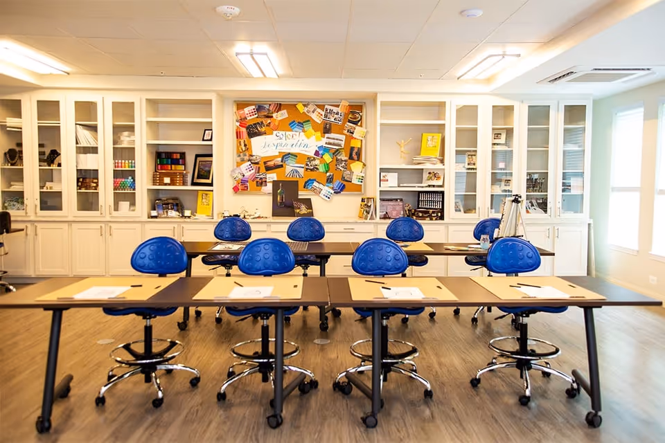 A bright and organized classroom or activity room with two rows of tables and blue ergonomic chairs on wheels. Each table has paper and writing materials placed on it. The back wall features white cabinets with glass doors displaying various supplies and decorations, and a colorful bulletin board with notes and pictures. The room has wood flooring and large windows letting in natural light.