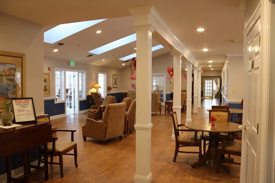 Interior view of a senior living facility common area with wooden floors, white pillars, and a mix of armchairs and dining tables with chairs. The space is well-lit with ceiling lights and natural light from skylights and windows. There are decorative elements such as paintings on the walls and small flower vases on the tables.