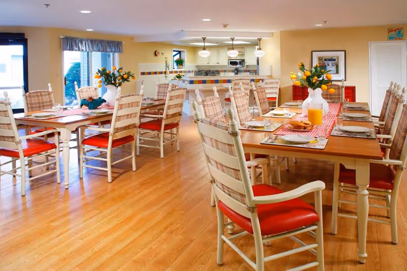 A bright dining room with wooden floors and multiple tables set for meals. Each table has white wooden chairs with red cushions and plaid backs. There are vases with orange flowers on the tables, and a pitcher of orange juice with plates of food. The room has large windows with curtains and a kitchen area visible in the background.