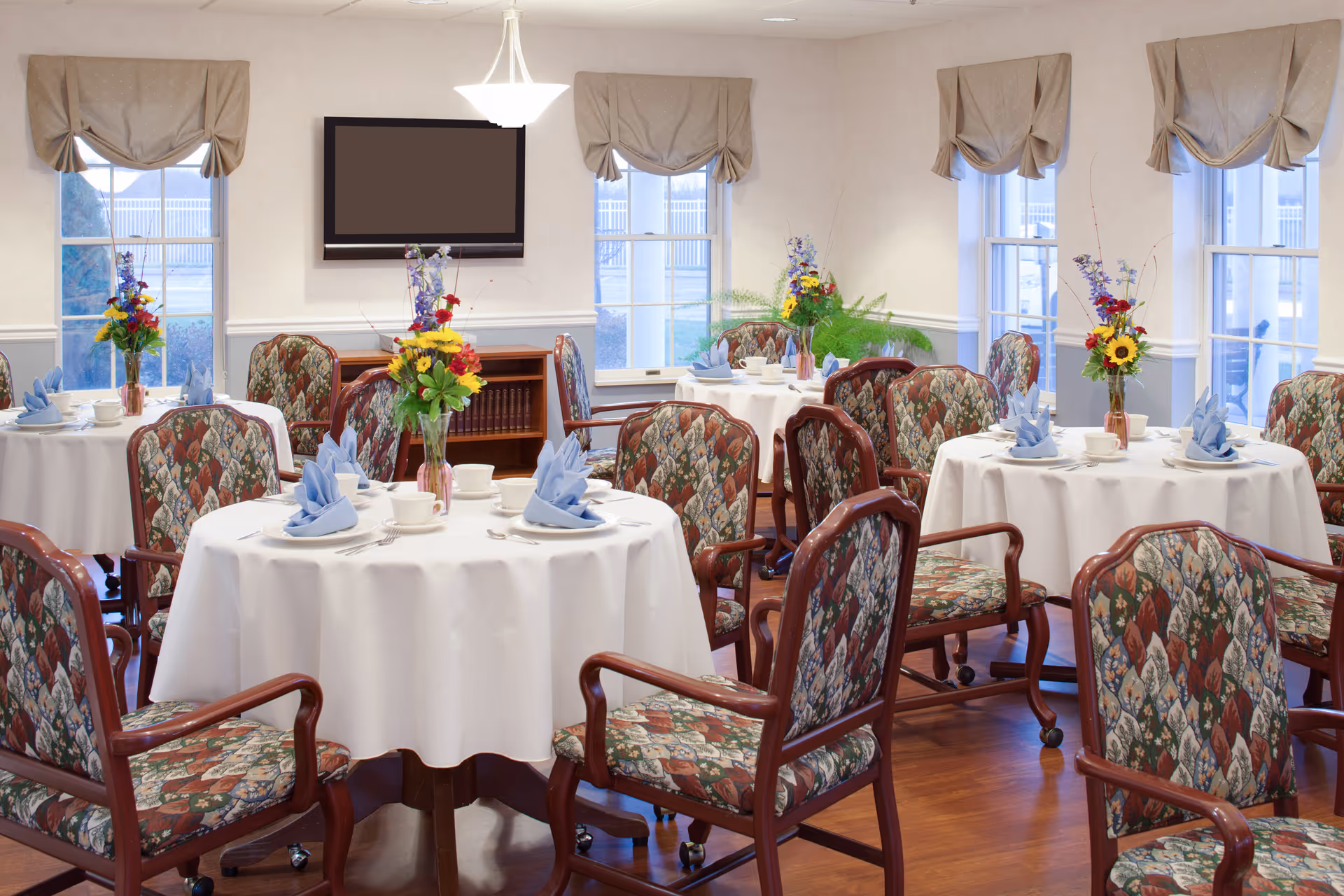 A dining room with round tables covered in white tablecloths, each set with blue folded napkins, white cups, and silverware. The chairs have wooden frames and patterned upholstery. There are flower arrangements in vases on each table. The room has several windows with beige valances, a wall-mounted flat screen TV, and a wooden cabinet with books underneath it.