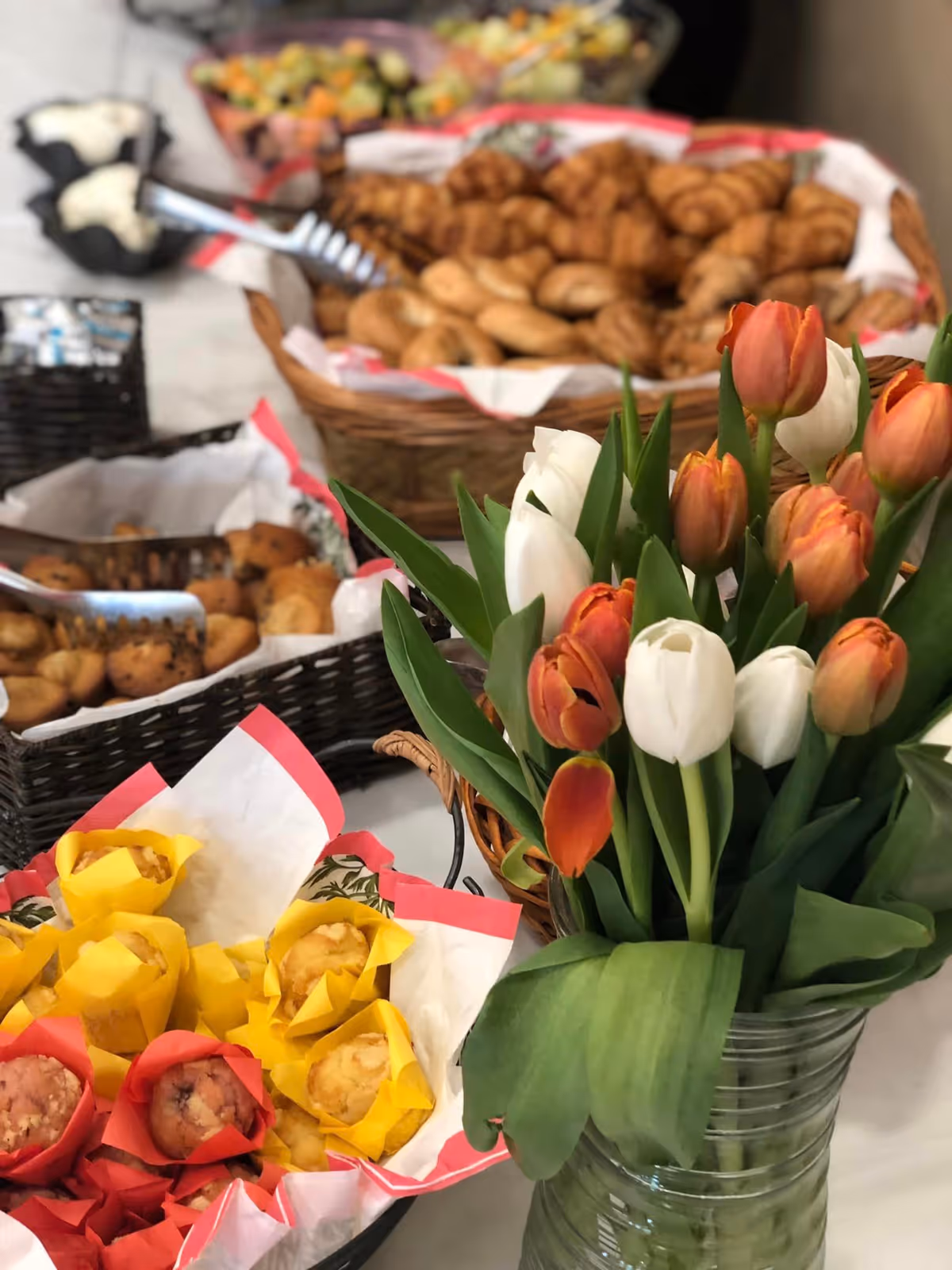 A close-up view of a table with baskets of assorted muffins and bagels, accompanied by a bouquet of orange and white tulips in a glass vase.