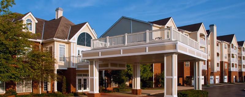 Exterior view of Edgewood Summit senior living facility showing a large covered entrance with white pillars and railings, multiple windows, and a multi-story building under a clear blue sky.