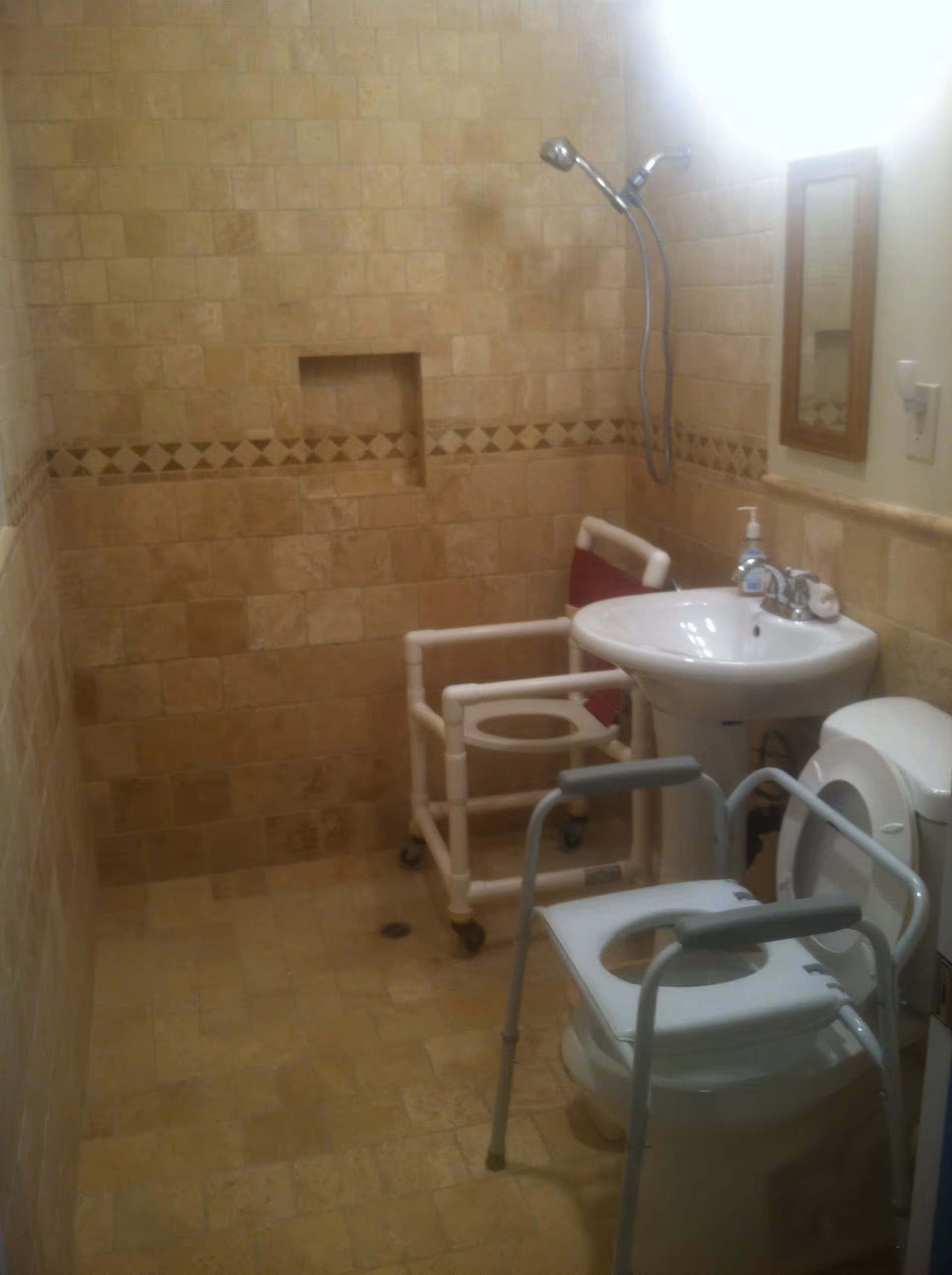 A bathroom with beige tiled walls and floor, featuring a shower area with a handheld showerhead, a white pedestal sink with a soap dispenser, a toilet with a raised seat and armrests, and a white PVC frame shower chair with wheels. There is a small rectangular mirror on the wall above the sink.