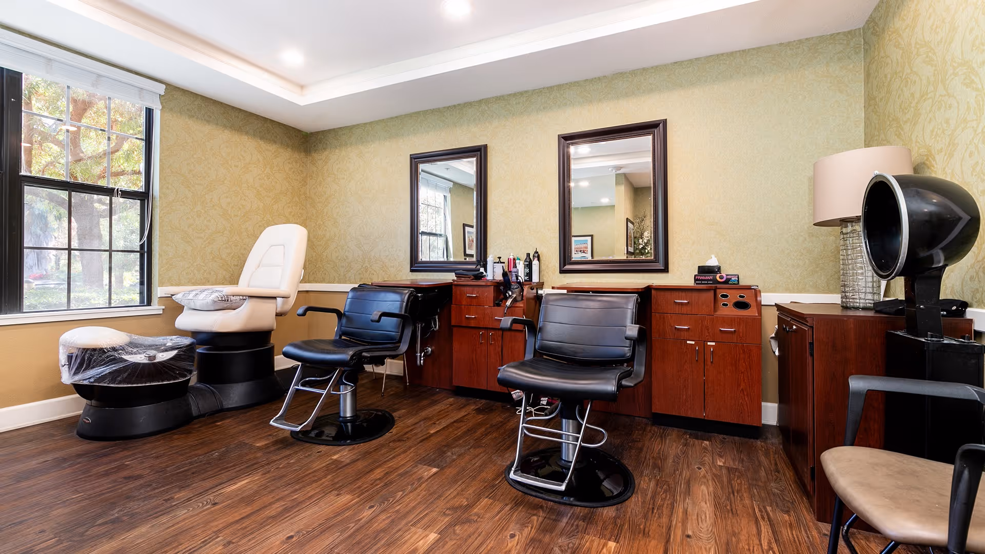 Interior view of a salon area in a senior living facility with two black salon chairs in front of mirrors mounted on a wall with wooden cabinets below. There is a white pedicure chair with a foot bath covered in plastic near a window with a view of trees outside. A hair dryer hood and a lamp are also visible on the right side of the room.