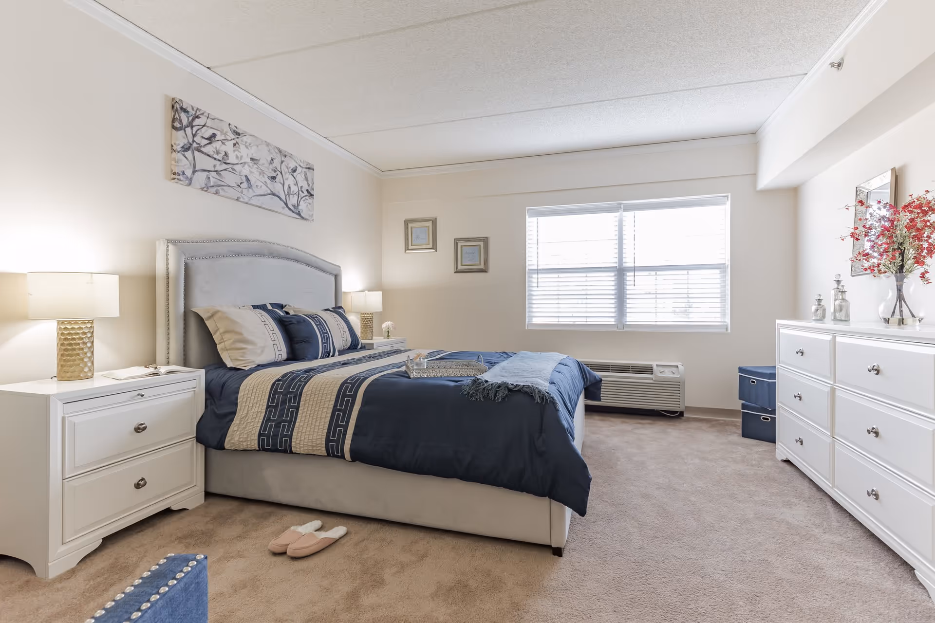 A neatly arranged bedroom with a large bed featuring blue and beige bedding, two bedside tables with lamps, a white dresser with decorative items and flowers, a window with blinds, and beige carpeted floor. There are slippers on the floor near the bed and framed artwork on the walls.