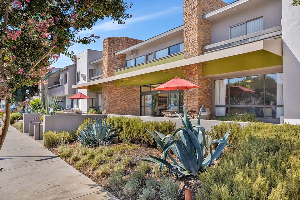 Exterior view of a modern senior living facility with a sidewalk, landscaped garden featuring agave plants and shrubs, and a building facade with large windows and red patio umbrellas providing shade to outdoor seating areas.