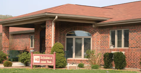 Exterior view of a single-story brick building with a red roof and a covered entrance. There are windows with shutters and some landscaping including bushes and a small tree. A sign near the entrance reads 'The Residence at Autumn Woods.'