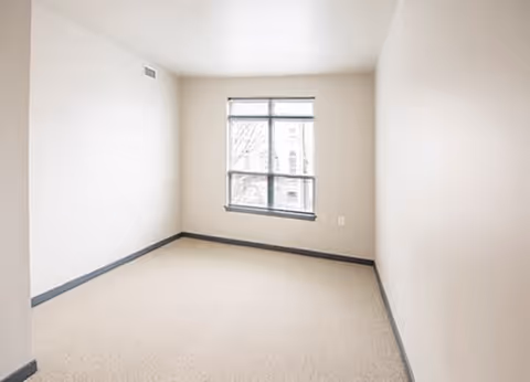 Empty small room with beige carpet, light-colored walls, a single window with blinds, and gray baseboards.