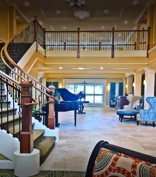 Spacious and well-lit interior common area featuring a grand piano near the center, a staircase with wooden handrails and carpeted steps on the left, and comfortable seating including armchairs and a cushioned bench. The area has tiled flooring, large windows with blue curtains, and a balcony railing overlooking the space from the upper level.