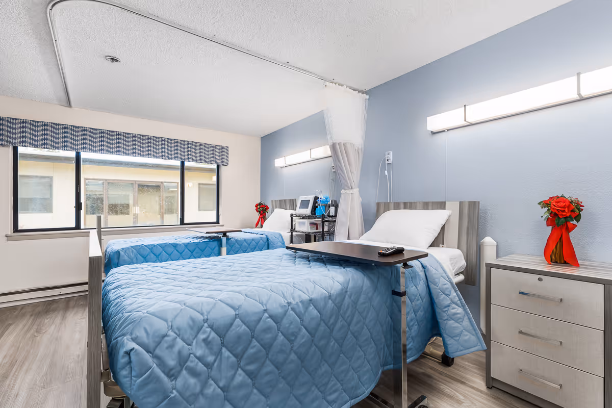 A bright and clean room with two hospital-style beds covered in light blue quilts. Each bed has a white pillow and an overbed table, one with a remote control on it. There is a small bedside cabinet with a vase of red flowers tied with a red ribbon. The room has a large window with a blue and white patterned valance, light blue walls, and wood-look flooring. Medical equipment is visible between the beds, and a privacy curtain is partially drawn.