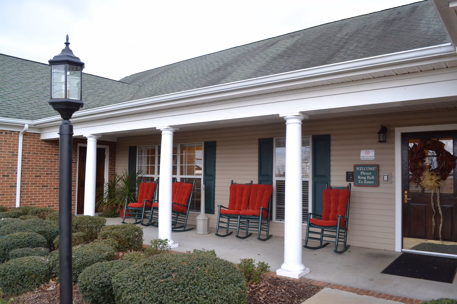 Front porch of a senior living facility with four red cushioned rocking chairs lined up against the wall under a covered area supported by white columns. There is a dark wooden door with a wreath and a sign that reads 'WELCOME! Please Ring Bell To Enter'. Bushes and a black lamp post are visible in the foreground.