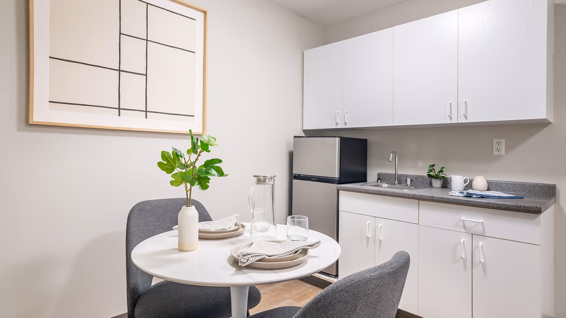 A small kitchenette area with white cabinets, a gray countertop, a stainless steel mini refrigerator, and a sink. In front of the kitchenette is a round white table set for two with plates, napkins, glasses, and a vase with green leaves. A modern abstract framed artwork hangs on the wall above the table.