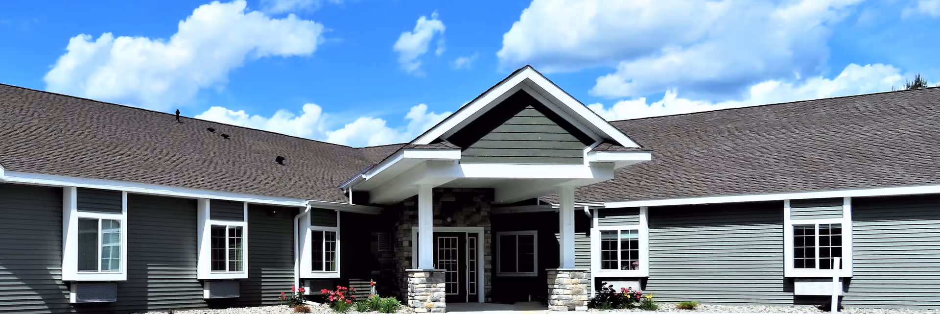 Exterior view of Coleman Fields Assisted Living & Memory Care building with gray siding, white trim, stone pillars at the entrance, and a clear blue sky with scattered clouds.