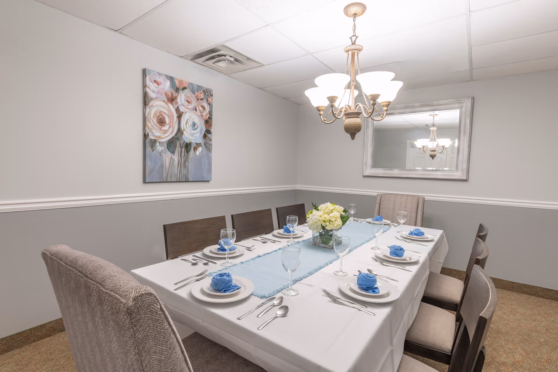 A dining room with a rectangular table set for six people. The table is covered with a white tablecloth and a light blue table runner, with white plates, silver cutlery, wine glasses, and blue napkins folded on each plate. There is a floral centerpiece with white flowers in the middle of the table. The room has light gray walls with a white chair rail, a floral painting on one wall, a large mirror on another wall, and a chandelier hanging above the table.