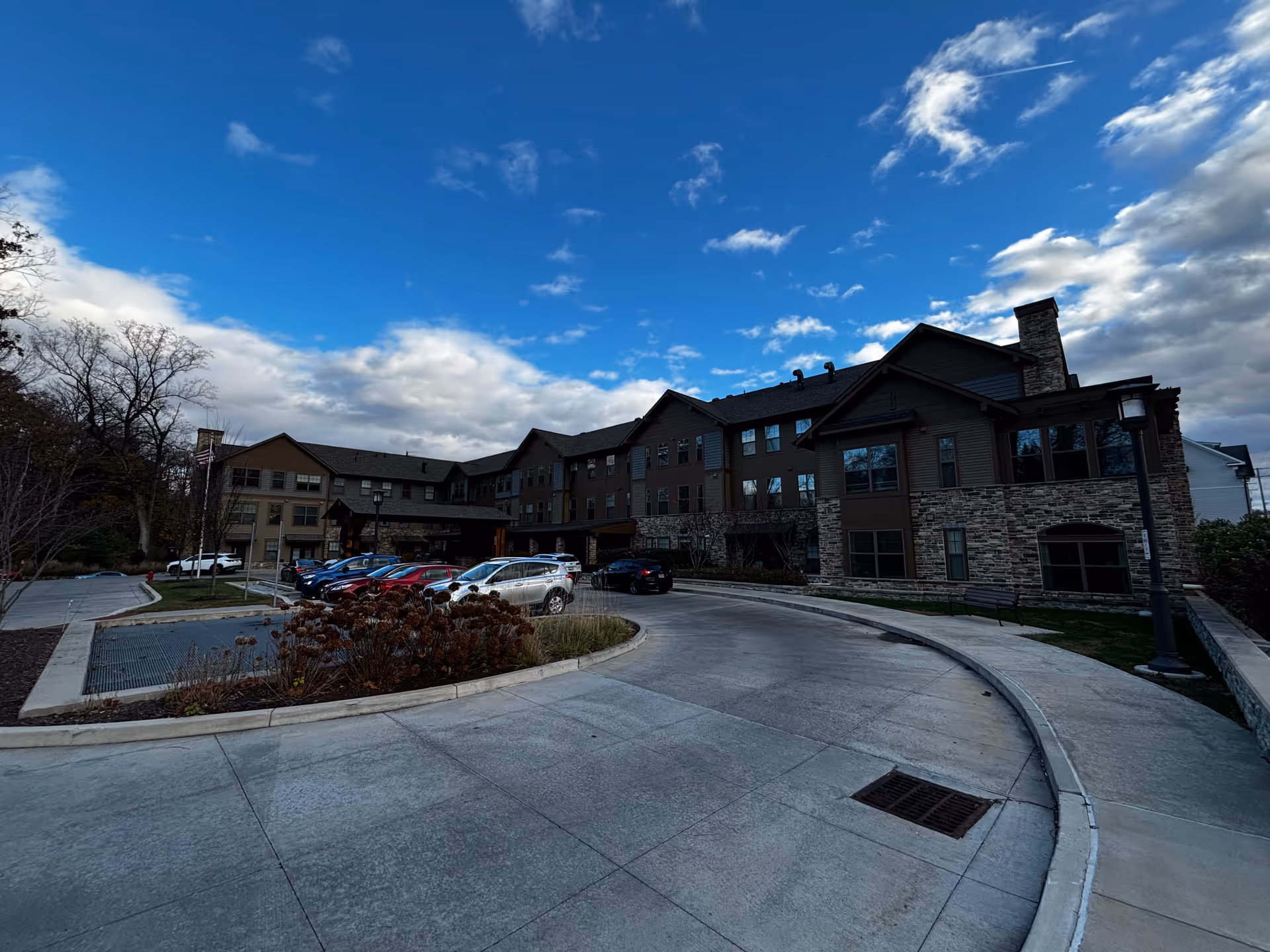 Front exterior of a multi-story senior living building with a curved driveway and parked cars under a blue sky.