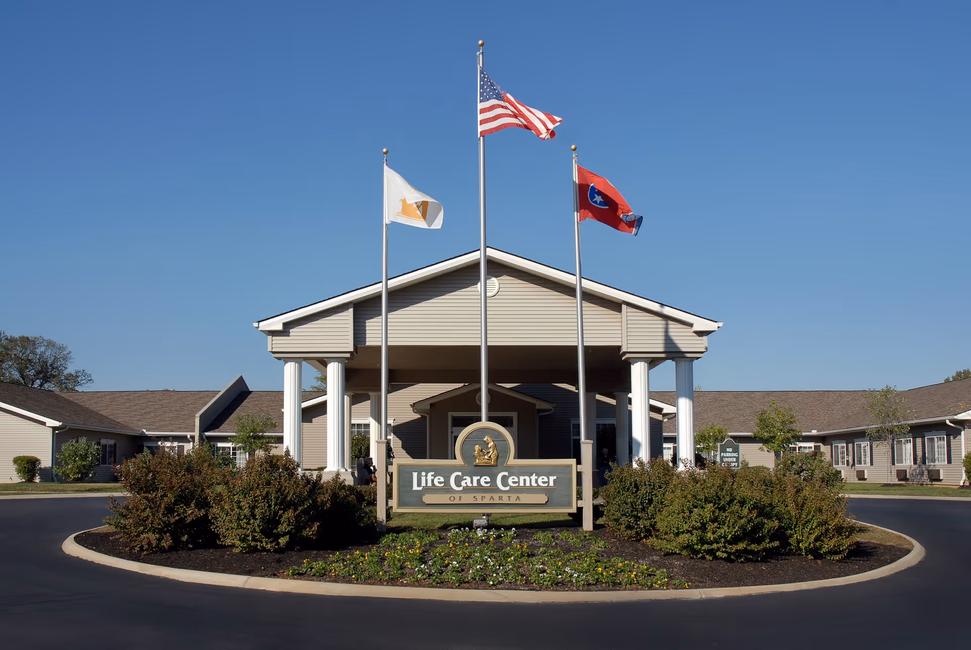 Front exterior view of the Life Care Center of Sparta building with three flagpoles displaying the American flag, a white flag, and a red flag. The building has a covered entrance with white columns and is surrounded by bushes and a circular driveway.