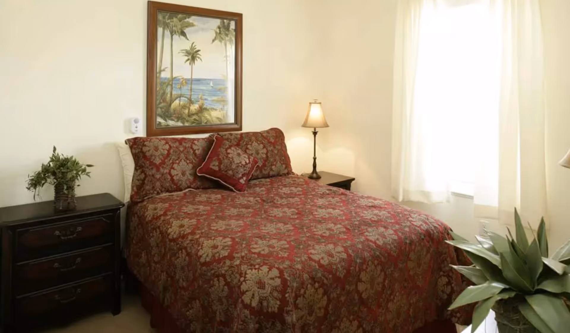 A bedroom with a bed covered in a red and gold patterned bedspread and matching pillows. There is a dark wooden nightstand with a plant on the left side of the bed and a lamp on a nightstand on the right side. A framed painting of a tropical beach scene hangs on the wall above the bed. Light filters in through a window with white curtains on the right side of the room. A large green plant is visible in the bottom right corner.