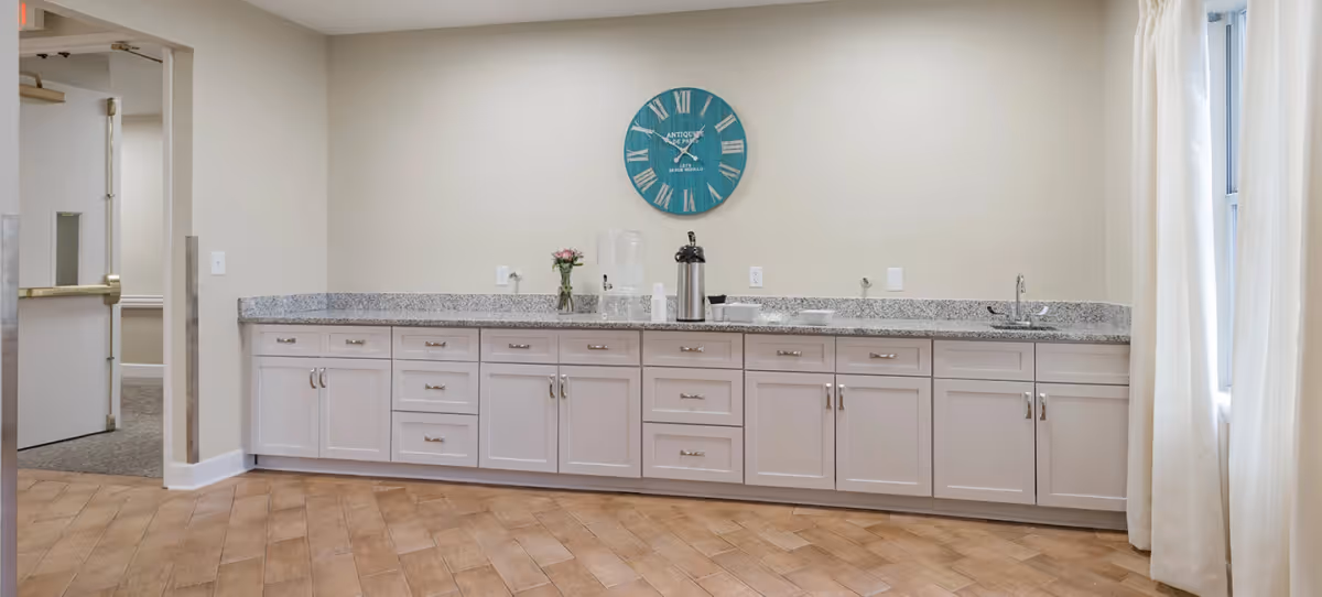 A long countertop with white cabinets underneath, a granite surface, a small sink on the right, a water dispenser, a coffee dispenser, cups, and a small vase with flowers. A large blue wall clock with Roman numerals hangs on the beige wall above the counter. The floor is tiled with a wood-like pattern, and there is a doorway on the left leading to another room.