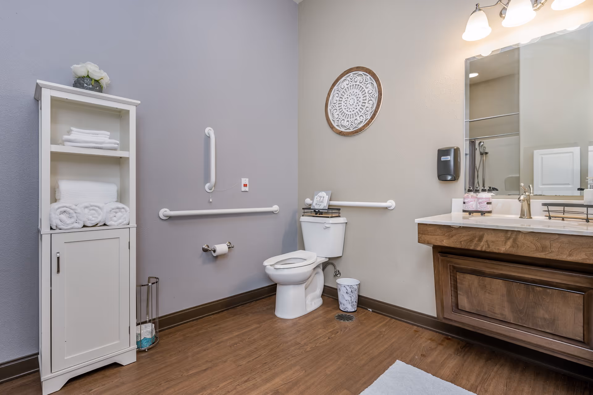 A clean and accessible bathroom with a white toilet equipped with grab bars on the walls for support. There is a white cabinet with neatly folded towels on the left side, a wooden vanity with a sink and mirror on the right, and a decorative round wall hanging above the toilet. The floor is wooden, and the walls are painted in light gray and beige tones.