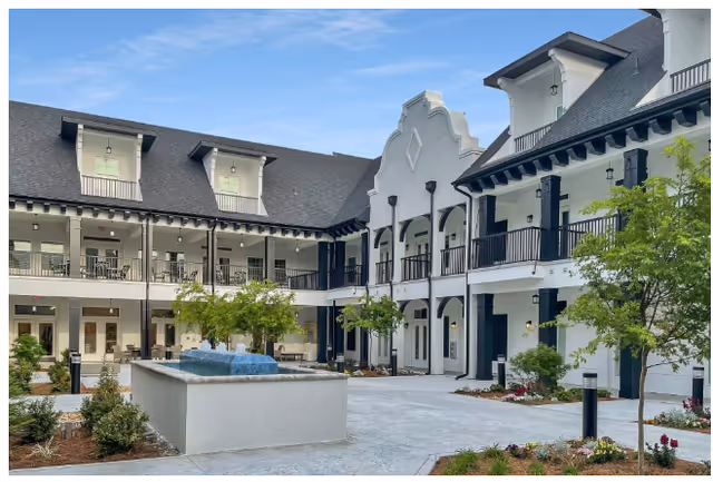 Outdoor courtyard area of The Blake at LPGA facility featuring a central water fountain, landscaped greenery, and a two-story building with balconies and large windows under a clear blue sky.