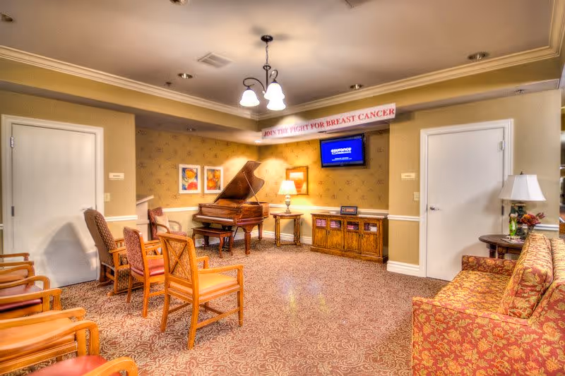 A cozy senior living common area with a grand piano, several wooden chairs with cushions, a patterned carpet, a floral upholstered sofa, a wooden cabinet with a lamp, and a wall-mounted TV. The walls are decorated with framed artwork and a banner that reads 'JOIN THE FIGHT FOR BREAST CANCER'.