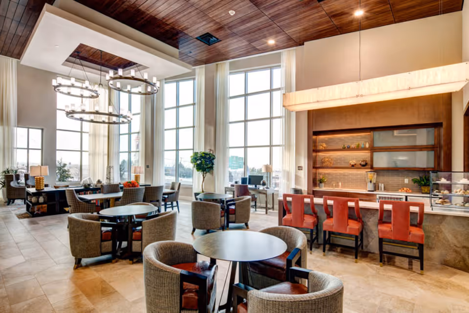 Bright communal dining and lounge area with round tables, upholstered chairs, tall windows and a bar counter with red stools.