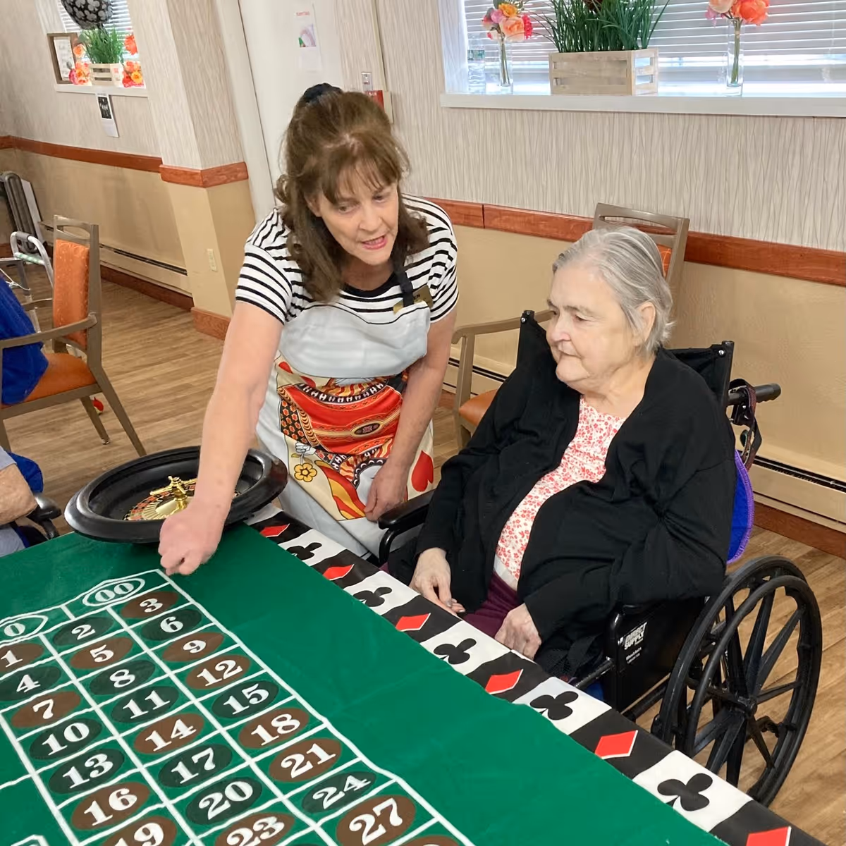 A woman in a striped shirt and apron is engaging with an elderly woman in a wheelchair at a table with a green felt roulette game layout. The setting appears to be a communal room with wooden flooring, chairs, and windows with blinds and flower decorations.