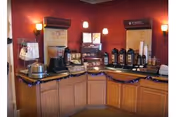 A coffee and beverage station with multiple coffee dispensers, a coffee maker, cups, lids, and condiments arranged on a wooden counter against a red wall with two wall-mounted light fixtures.