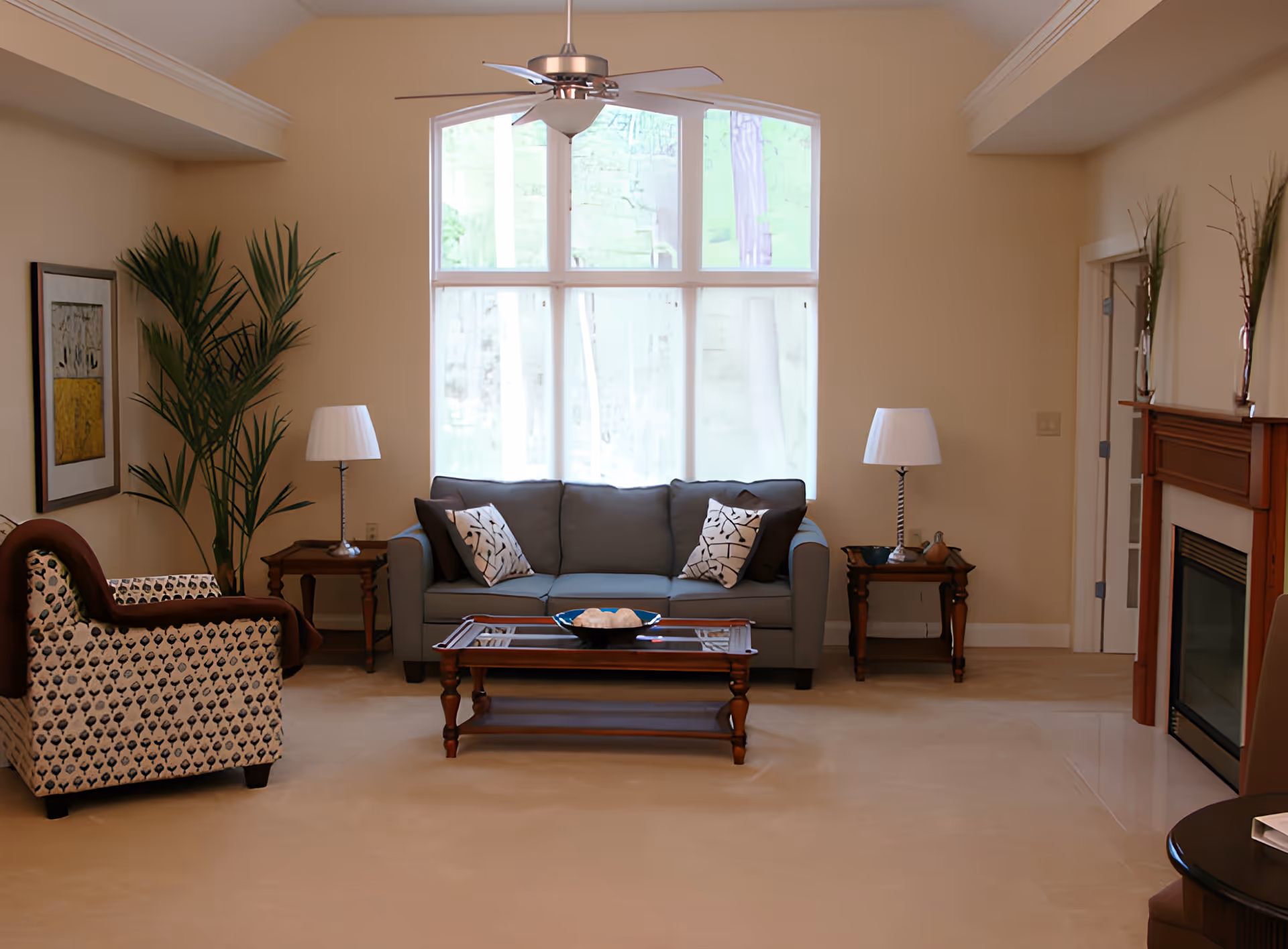 Well-lit living room with a sofa, patterned armchair, coffee table, lamps, and a fireplace beneath a large arched window.