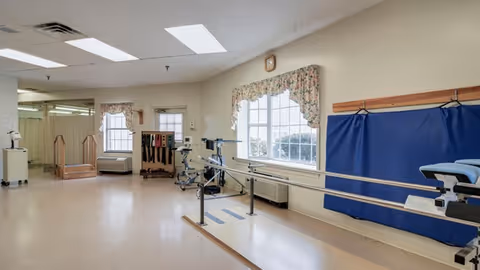 A spacious rehabilitation room with parallel bars for walking exercises, a padded treatment table, and various therapy equipment. The room has large windows with floral curtains allowing natural light, beige walls, and a clean, open floor space.