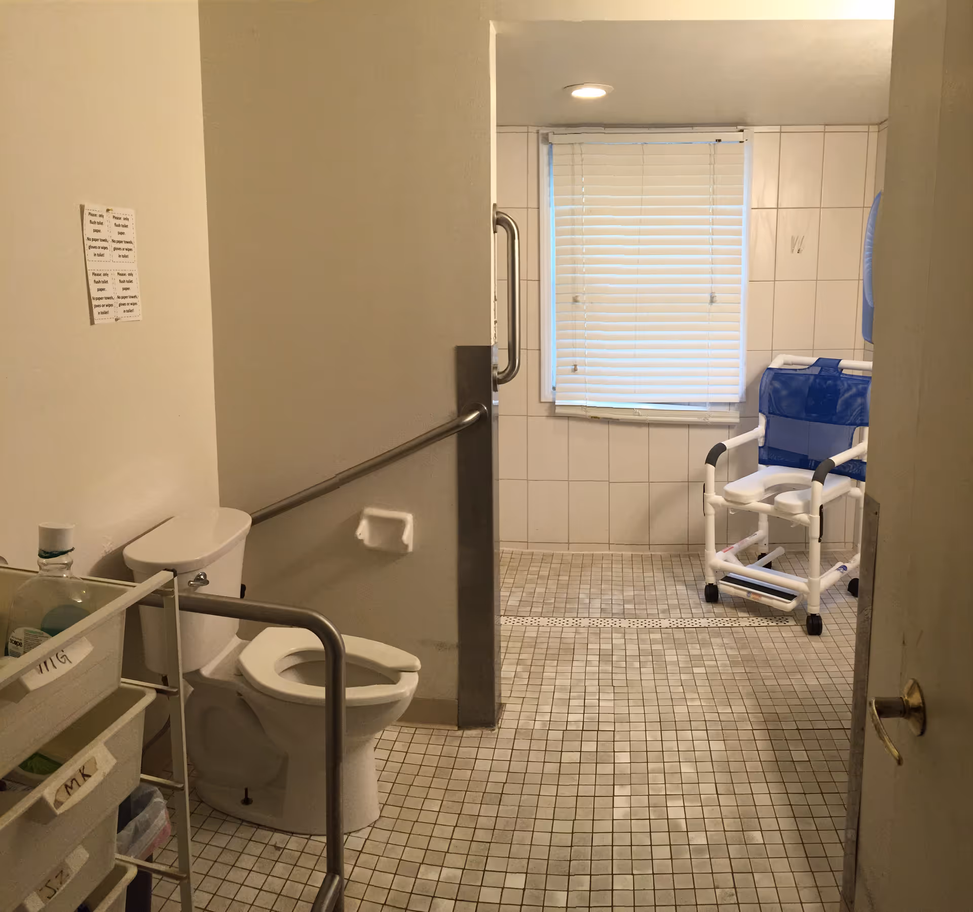 Accessible bathroom with a toilet, grab bars, tiled floor and a shower chair near a window with blinds.