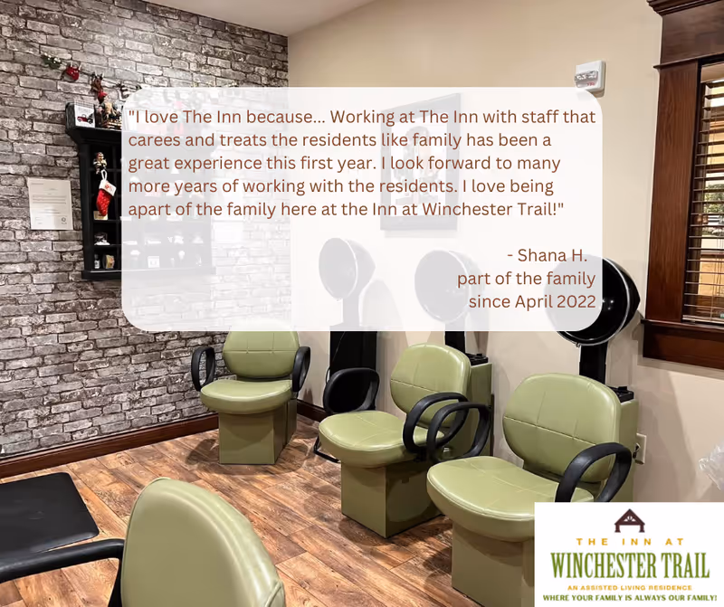 Interior view of a salon area in The Inn at Winchester Trail featuring four green salon chairs with black armrests, three black hair dryers mounted on stands, a brick accent wall with shelves holding decorative items, and a window with wooden blinds. There is a testimonial text overlay from Shana H. about working at the facility, and a logo for The Inn at Winchester Trail in the bottom right corner.