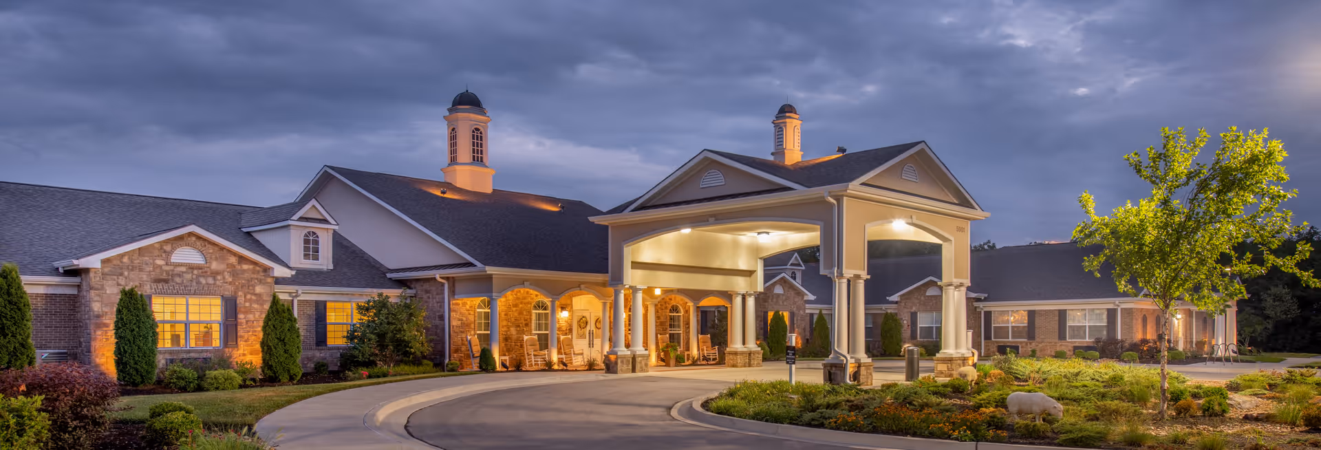 Exterior view of Benton House of Tiffany Springs senior living facility at dusk, showing a large building with stone and brick facade, a covered entrance with columns, well-maintained landscaping, and illuminated windows.