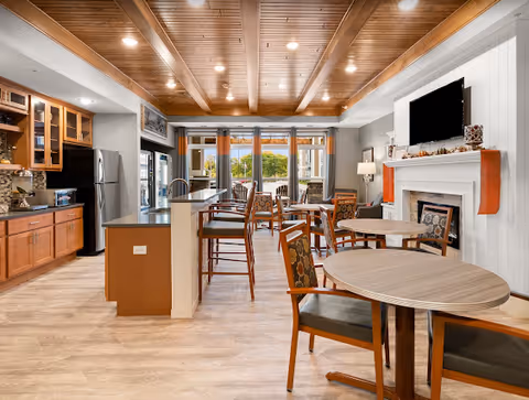 A bright and spacious common area in a senior living facility featuring a kitchen with wooden cabinets and stainless steel refrigerator on the left, a breakfast bar with high chairs, multiple round tables with cushioned chairs, a fireplace with a mounted flat-screen TV above it, and large windows with curtains letting in natural light.