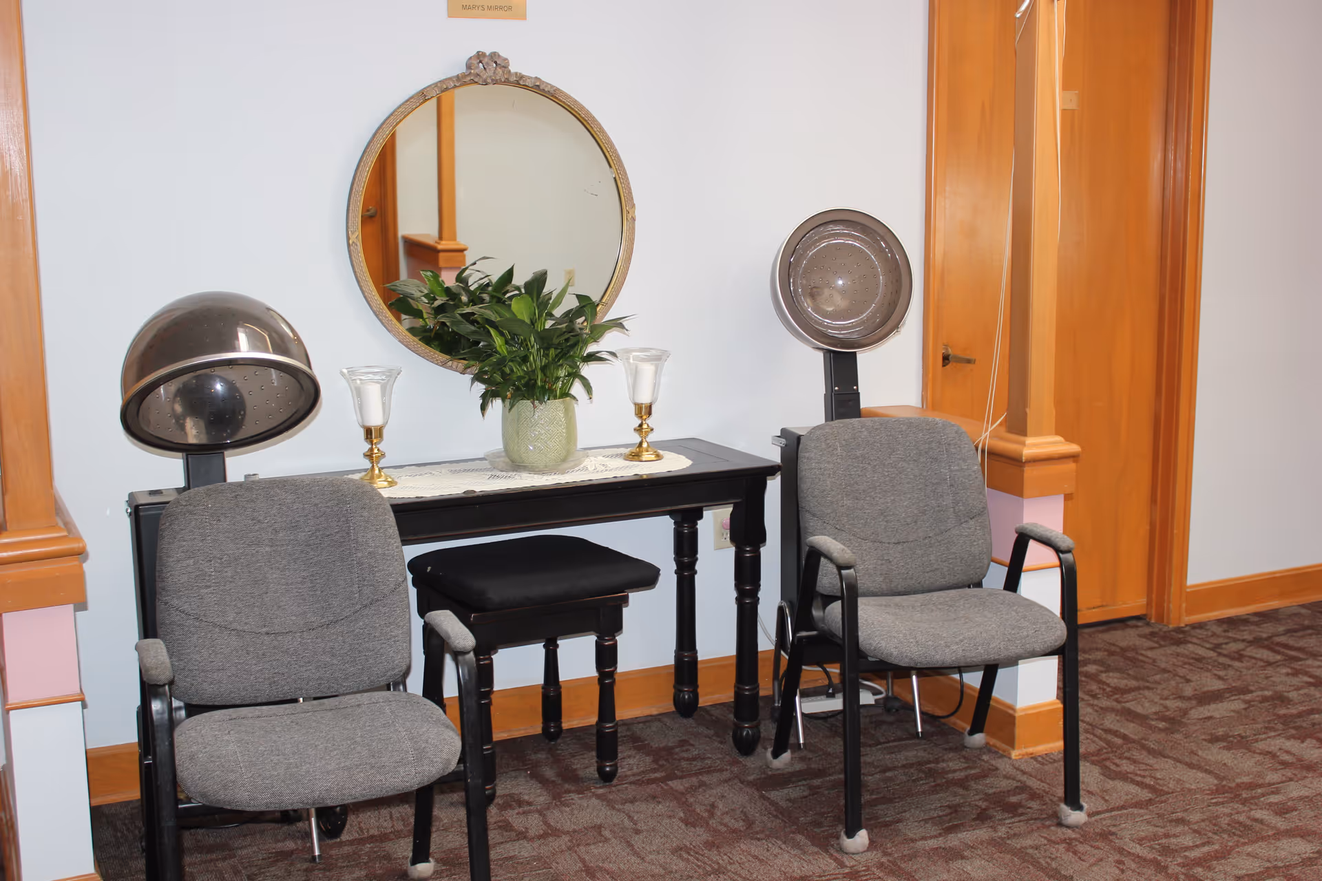 A small salon area with two gray chairs positioned under two vintage hair dryers. Between the chairs is a black table with a green potted plant and two candle holders. Above the table is a round mirror labeled 'Mary's Mirror'. The room has light blue walls, wooden trim, and a carpeted floor.