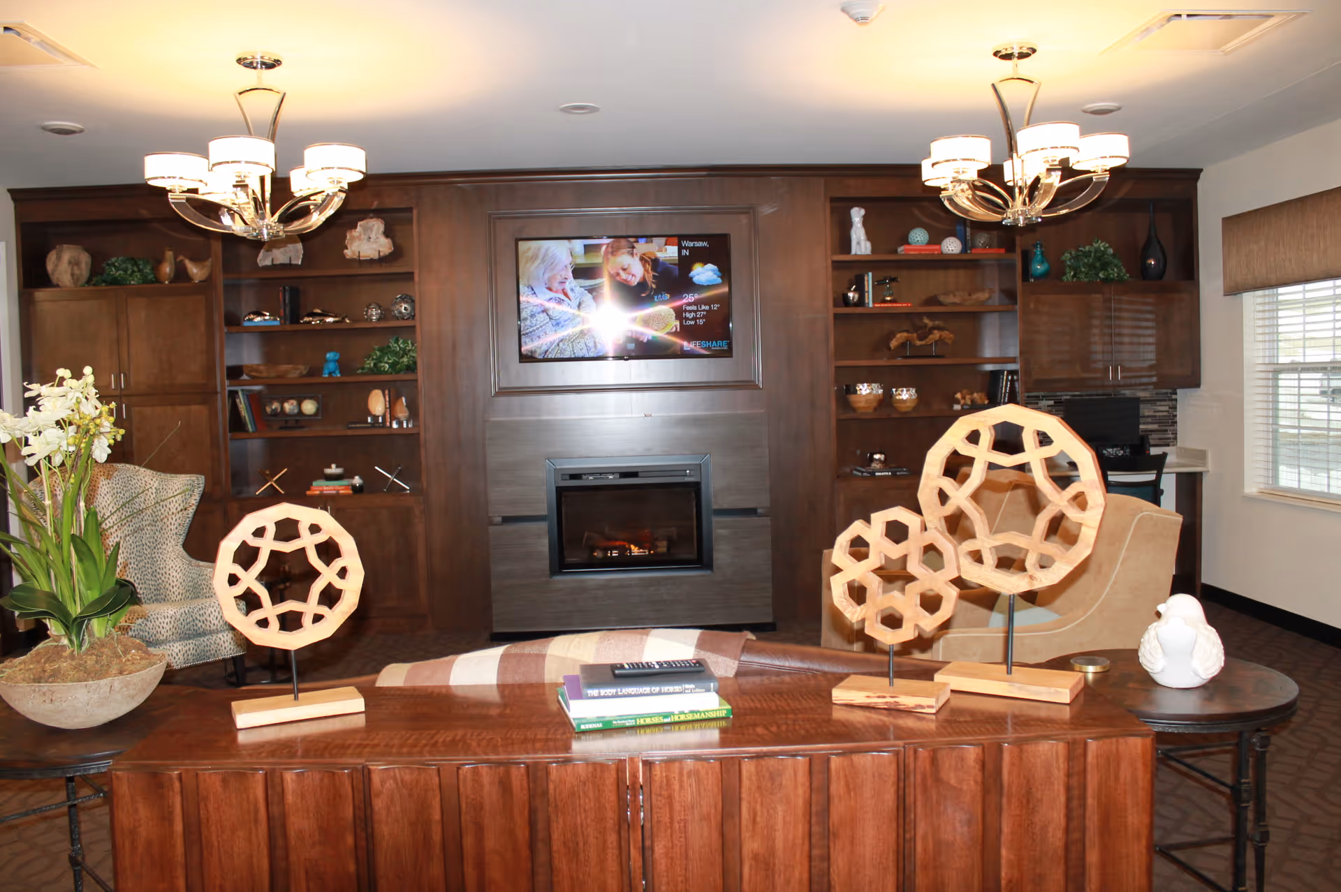 A cozy common lounge with built-in wooden shelving and a fireplace topped by a wall-mounted TV, chandeliers, seating, and decorative wooden sculptures on a console table.