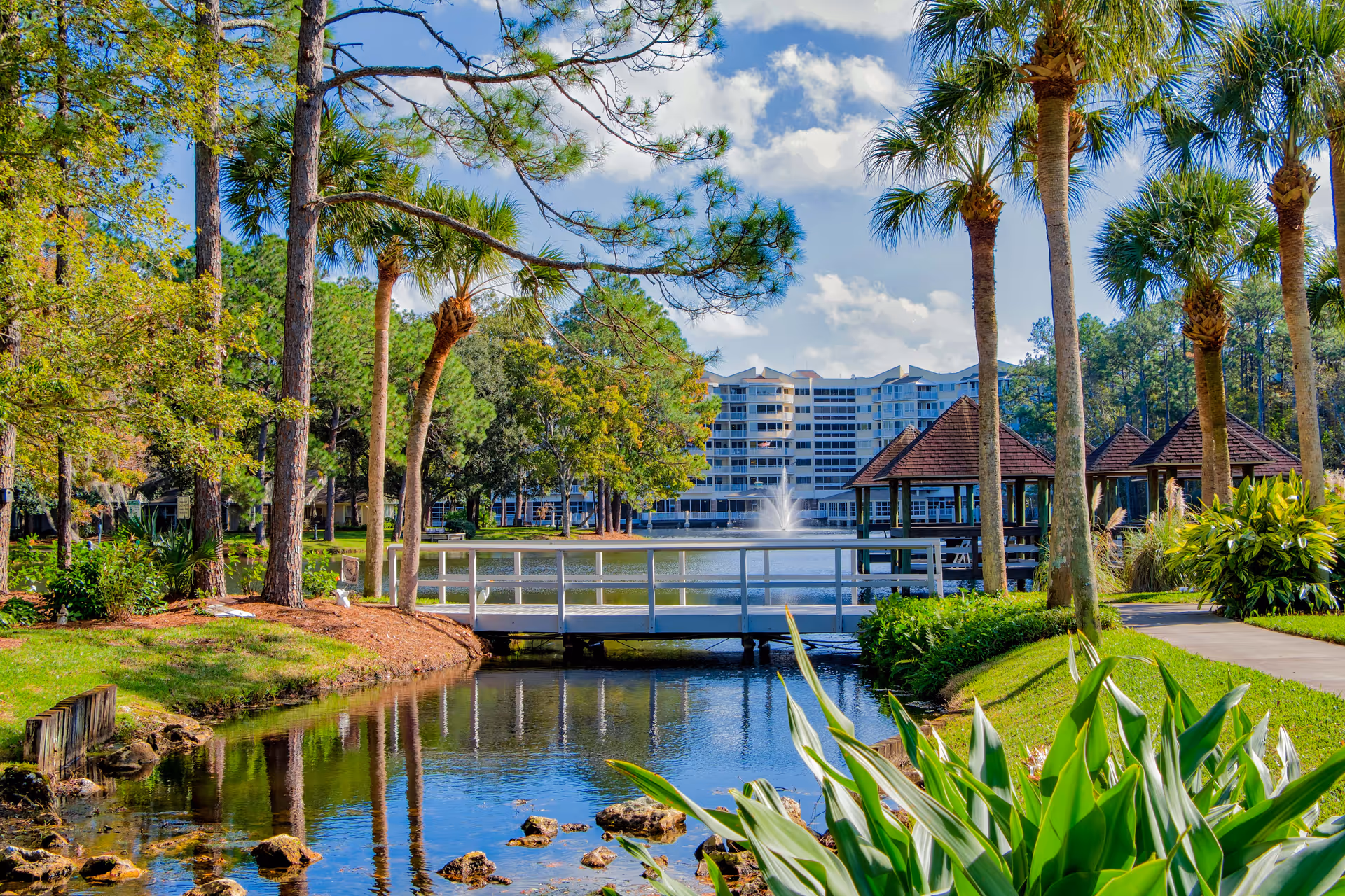 A landscaped pond with a white footbridge, palm trees, gazebos, and a multi-story building with a fountain in the background.