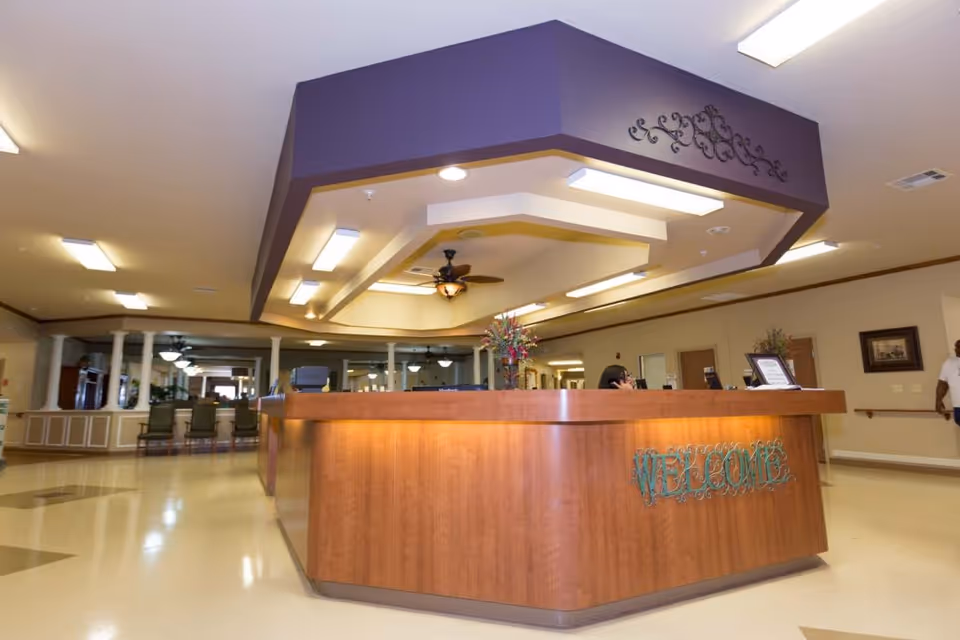 Interior view of a healthcare facility reception area with a large wooden welcome desk, ceiling fan, decorative lighting, and seating area in the background. A person is partially visible behind the desk and another person is walking in the hallway.